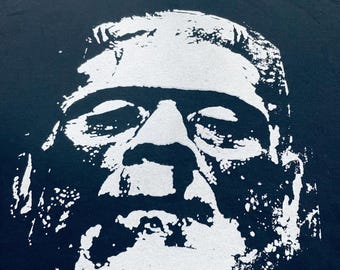 May include: A black crew neck t-shirt featuring a large, high-contrast white graphic print of a stylised face with heavy brow ridges, a flat top, and stitched details, rendered in a distressed, ink-stamped aesthetic on the front chest.