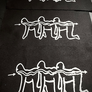 Op de afbeelding: Drie zwarte stoffen patches met witte lijntekeningen van mensen die hand in hand lopen in een rij. De mensen zijn getekend in een eenvoudige, cartoonachtige stijl. De patches zijn aan elkaar genaaid, waarbij de bovenste patch de middelste patch lichtjes overlapt en de middelste patch de onderste patch lichtjes overlapt.