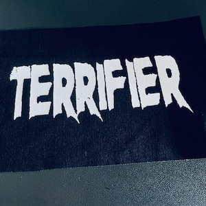 Può includere: Toppa nera con la parola "TERRIFIER" in bianco, in lettere stilizzate.