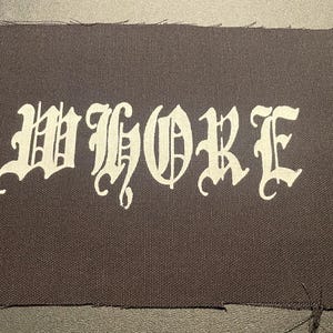 May include: A black fabric patch with the word "WHORE" written in white, stylized lettering.