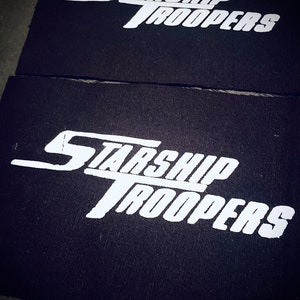 Könnte beinhalten: Zwei schwarze Stoff-Patches mit weißer Aufschrift "Starship Troopers".