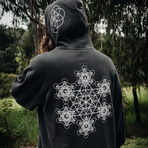 Puede incluir: Una sudadera con capucha negra con un diseño geométrico blanco en la espalda. El diseño es un patrón complejo de círculos y líneas, que se asemeja a una flor o un mandala.