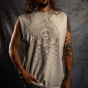 May include: An off-white sleeveless t-shirt with a brown geometric mandala design. The shirt is worn by a person with long hair and tattoos on their arm. The person is wearing a gold bracelet and grey jeans. The background is a mottled grey.