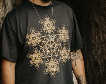 May include: Black t-shirt with a gold geometric design and a silver pendant necklace. The design features interconnected shapes. The person in the image is wearing a black hat.
