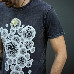 Puede incluir: Camiseta gris oscuro con un diseño de esferas geométricas blancas. Un colgante dorado cuelga de un cordón negro. La camiseta tiene un aspecto descolorido y texturizado, y el diseño presenta esferas interconectadas.