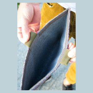 May include: Open view of a felt laptop sleeve with olive green fabric handles. The interior is a dark gray color, and the exterior has a multicolored tweed pattern. The sleeve is being held open, showcasing its capacity.