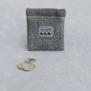 May include: A small, gray tweed coin purse with a rectangular shape and a snap closure. The purse has a leather patch with the word "bright" and three metal studs. Several coins are scattered in front of the purse.