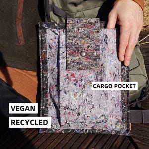 Op de afbeelding: Een rechthoekige, veganistische, gerecyclede tas met een cargozak. De tas is gemaakt van een grijs, veelkleurig materiaal met rode, blauwe en zwarte accenten. De tas heeft een klepsluiting met een zilveren drukknop. De tekst "VEGAN RECYCLED" is zichtbaar.
