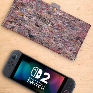 Può includere: Una console Nintendo Switch grigia con schermo colorato, accanto a una custodia in feltro multicolore. La custodia ha una chiusura a linguetta grigia con un bottone dorato. Lo schermo della Switch mostra "Nintendo Switch".