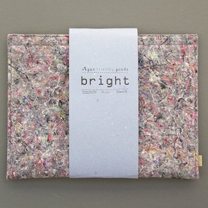 May include: A gray laptop sleeve with a colorful paint splatter design. The sleeve has a light gray paper band with the words "vegan friendly goods" and the word "bright" printed on it. A small yellow tag is attached to the side.