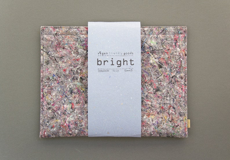 May include: A gray laptop sleeve with a colorful paint splatter design. The sleeve has a light gray paper band with the words "vegan friendly goods" and the word "bright" printed on it. A small yellow tag is attached to the side.