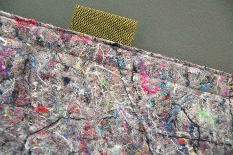 May include: Close-up of a gray, multi-colored fabric with a textured appearance. The fabric is composed of interwoven fibers in shades of gray, with vibrant flecks of pink, blue, green, and orange. A rectangular olive-green strap is attached to the fabric.