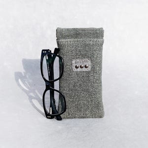 May include: Black eyeglasses with a gray fabric case. The case has a leather tag with the word "bright" and three small silver rivets. The glasses are next to the case on a light gray surface.