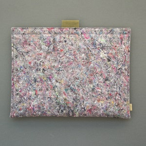 May include: A rectangular tablet sleeve with a colorful, abstract pattern. The sleeve is primarily gray with a mix of red, blue, and white accents. It has a small, tan fabric loop and a small tan tag.
