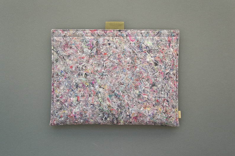 May include: A rectangular tablet sleeve with a colorful, abstract pattern. The sleeve is primarily gray with a mix of red, blue, and white accents. It has a small, tan fabric loop and a small tan tag.