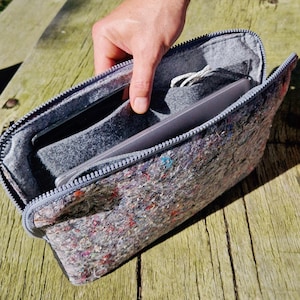 May include: A gray zippered laptop sleeve made of a speckled, felt-like material. The interior is gray and holds a laptop and other items. The zipper is gray, and the sleeve is open, revealing the contents. The sleeve is on a wooden surface.