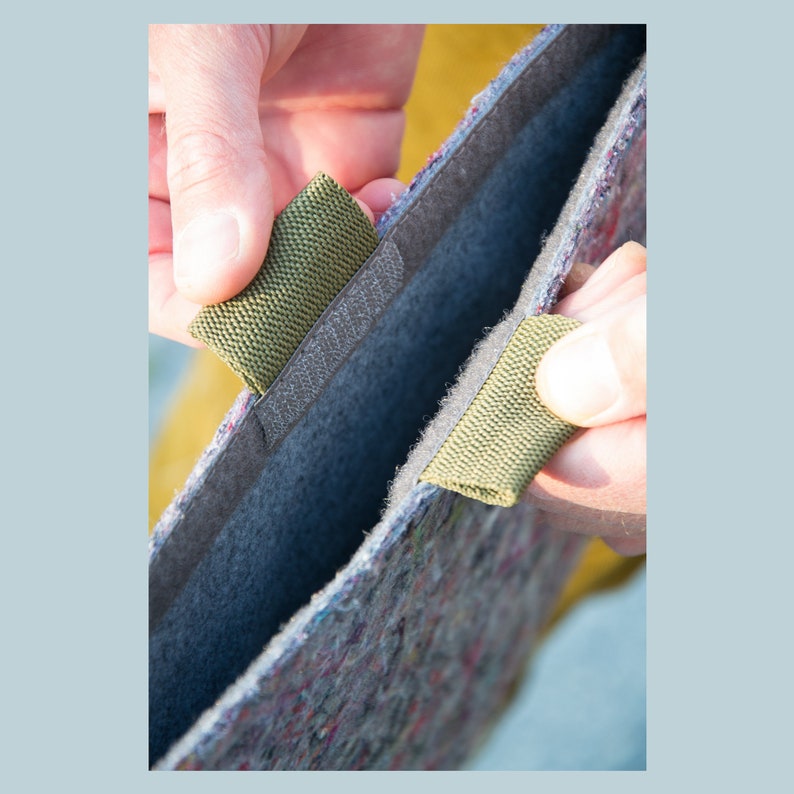 May include: Close-up of a felt bag with olive green fabric tabs and a velcro closure. The bag is made of a multi-colored, speckled material with a gray interior. The tabs are being held open to show the inside of the bag.