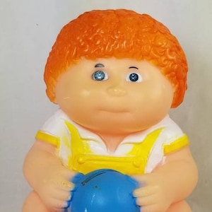 May include: A plastic Cabbage Patch Kid doll with orange hair, wearing a yellow and white outfit, holding a blue ball.