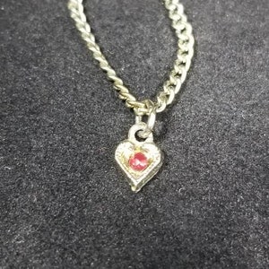 May include: A silver chain necklace with a heart-shaped pendant. The pendant has a red gemstone in the center.
