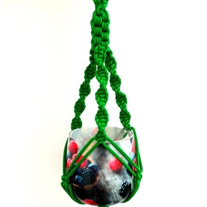 Puede incluir: Un macetero de macramé verde con una maceta de vidrio. La maceta tiene un diseño de bayas.
