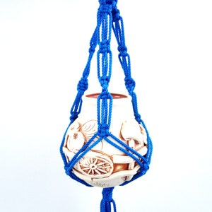 Puede incluir: Un macetero de macramé azul con una maceta de cerámica blanca que presenta un diseño detallado de un carro con ruedas y fruta.