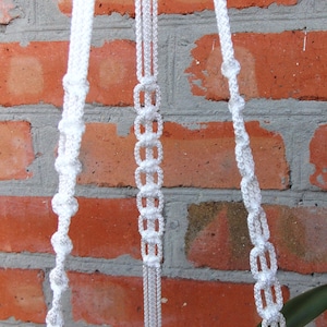Puede incluir: Un macetero de macramé blanco con tres hebras de cuerda, colgado de una pared de ladrillo rojo.