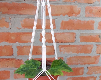 Pode incluir: Um suporte de planta de macramé branco com um vaso de terracota e uma planta verde com folhas penduradas em uma parede de tijolos.