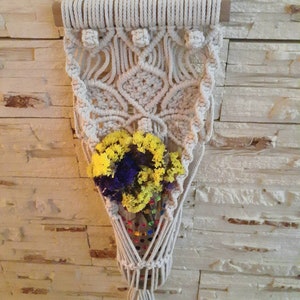Puede incluir: Un macramé de pared con una barra de madera en la parte superior. El macramé está hecho de cordón de algodón de color natural y tiene un patrón geométrico. Un pequeño ramo de flores secas está en el bolsillo del macramé.