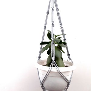 Peut inclure: Un porte-plante en macramé gris avec un pot blanc et une plante verte à l'intérieur. Le porte-plante est fait de cordon de coton et a un pompon en bas.
