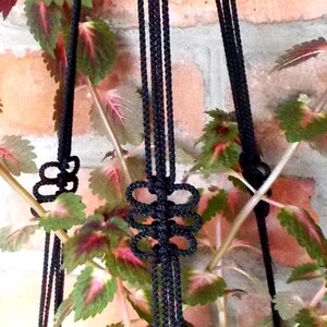 Puede incluir: Un macramé para plantas negro con un patrón de nudos. El macramé está hecho de cuerda gruesa y tiene tres hebras.