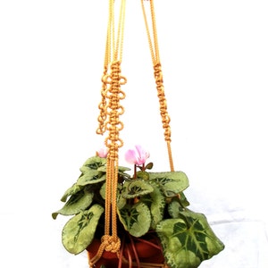 May include: A macrame plant hanger made of natural beige cord with a potted plant with green leaves and pink flowers hanging from it.