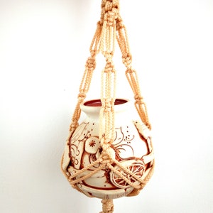 Puede incluir: Un macetero de macramé beige con una maceta de cerámica blanca con un diseño floral marrón. La maceta cuelga del macetero de macramé.