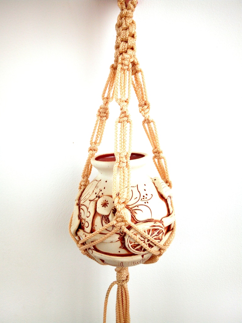 May include: A beige macrame plant hanger with a white ceramic pot featuring a brown floral design. The pot is suspended from the macrame hanger.
