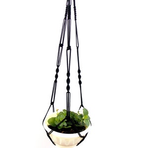 May include: Black macrame plant hanger with a white pot and green plant hanging from it.
