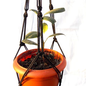 Puede incluir: Un macetero de macramé negro con una maceta de terracota y una pequeña planta verde en su interior. El macetero está hecho de cuerda gruesa y tiene un nudo en la parte superior para colgarlo.
