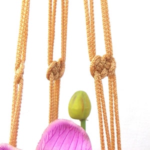 May include: A close-up of a macrame plant hanger with a light brown color. The hanger is made of multiple strands of rope that are knotted together. The hanger is hanging from the top of the image and there is a pink orchid flower at the bottom of the image.