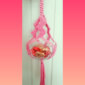 Puede incluir: Un macramé para colgar plantas de color rosa con un cuenco de vidrio lleno de chocolates envueltos en papel de aluminio dorado y rojo.