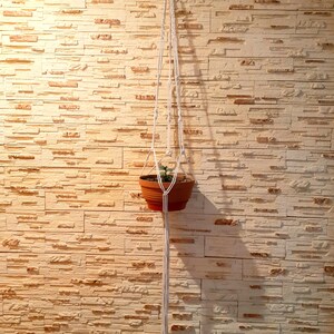 May include: A white macrame plant hanger with a terracotta pot and a small green plant hanging from it against a light beige brick wall.