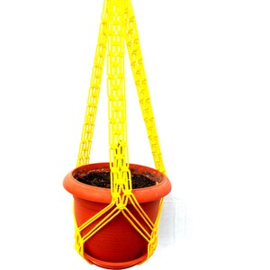 May include: A yellow macrame plant hanger with a terracotta pot. The hanger is made of thick, knotted rope and has a tassel at the bottom.