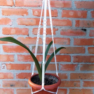 May include: A white macrame plant hanger with a brown terracotta pot and a green plant hanging from a brick wall.