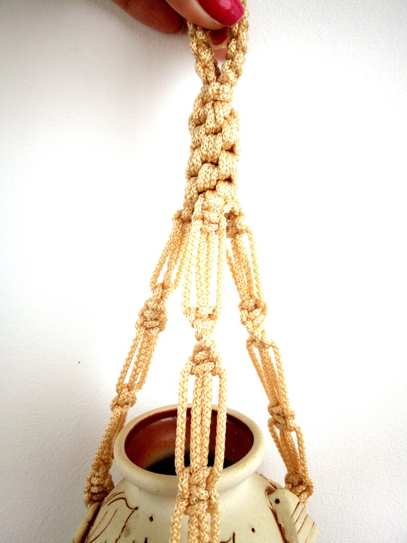 May include: A macrame plant hanger made with cream-coloured cord. The hanger is hanging from a ceramic pot with a brown rim.