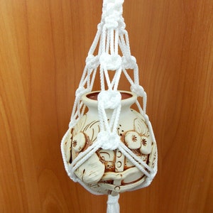 Puede incluir: Un macetero de macramé blanco con una maceta de cerámica blanca con un diseño floral marrón.