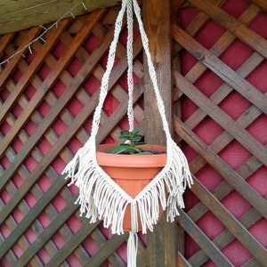 May include: A white macrame plant hanger with a brown pot and a green plant hanging from a wooden lattice.