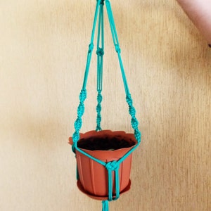 Puede incluir: Un macetero de macramé de color turquesa con una maceta de terracota. El macetero está hecho de cuerda anudada y tiene una borla decorativa en la parte inferior.
