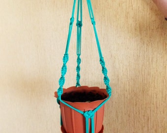 Pode incluir: Um suporte de plantas de macramé turquesa com um vaso de terracota. O suporte é feito de corda de nó e tem uma borla decorativa na parte inferior.