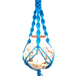 Puede incluir: Un macetero de macramé azul con una maceta de cerámica blanca. La maceta tiene un diseño floral.