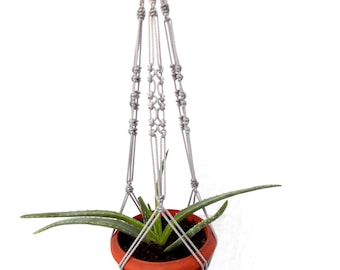 Pode incluir: Um suporte de plantas de macramé cinza com um pequeno vaso de terracota e uma planta suculenta verde dentro.