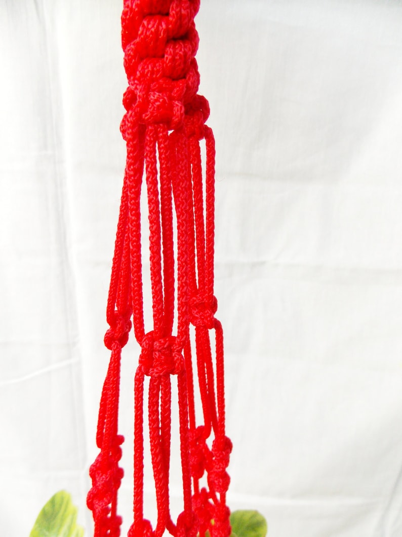K&ouml;nnte beinhalten: Ein roter Makramee-Pflanzenh&auml;nger mit einem geknoteten Design. Der H&auml;nger besteht aus dickem, rotem Seil und hat eine Reihe von Schlaufen, die eine netzartige Struktur bilden.