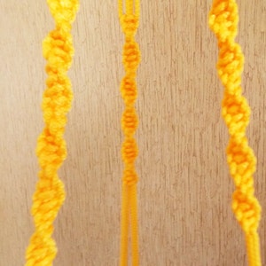May include: Three strands of yellow macrame cord hanging against a wood background.