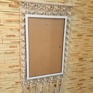 Puede incluir: Un tapiz de macramé blanco con un marco blanco y un inserto marrón. El tapiz tiene una franja de flecos en la parte inferior.
